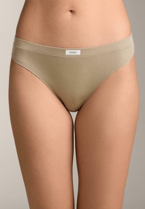 Bež bezešve spodnje perilo v bikini stilu z belem osrednjim etiketo na pasu, ki ga nosi oseba na nevtralnem ozadju.