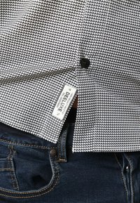 Overhemd met korte mouwen met een zwart-wit herhalend patroon, voorzien van een knoopsluiting en een zichtbaar etiket met de tekst "DEELUXE." Gecombineerd met donkere jeans.