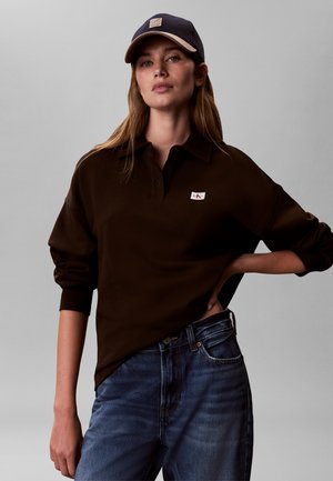 Kvinne iført mørkebrun langermet poloshirt, blå jeans og svart baseballcap, stående med en hånd på hoften mot ensfarget bakgrunn.