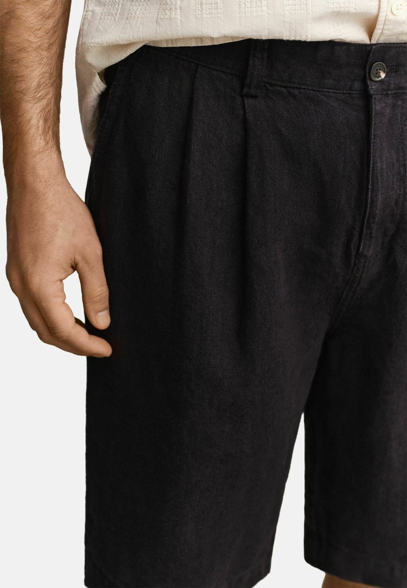 Plano detalle de una persona que lleva pantalones cortos plisados negros y una camisa de manga corta beige con textura, con la mano izquierda relajada a un lado.