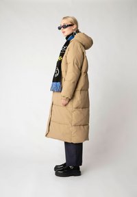 Long beige puffer coat with a hood, featuring horizontal stitching and elastic cuffs. Paired with a black scarf with blue fringe and black platform shoes.