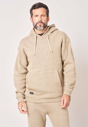 Man met baard die een beige hoodie en bijpassende broek draagt, staande voor een effen lichte achtergrond.
