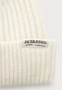 Crèmekleurige ribgebreide beanie met omgeslagen manchette en een wit label met de tekst "JACK&JONES STUDIO Collectie" dat op de voorkant is genaaid.