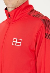 Röd zip-jacka med svarta mönstrade axelaccenter och en broderad flagga i vitt och rött. Tyget verkar slätt och har en atletisk design.
