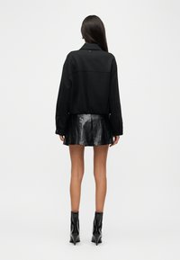 Μαύρο φαρδύ σακάκι με δομημένο γιακά, συνδυασμένο με μαύρη πλισέ φούστα από συνθετικό δέρμα. Το μοντέλο φοράει κομψά μαύρα μποτάκια αστραγάλου.