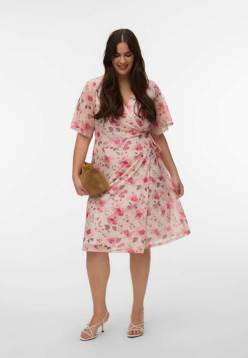 Femme portant une robe portefeuille rose à fleurs, tenant une pochette en daim marron, chaussée de talons blancs à lanières, debout devant un fond blanc uni.