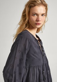 Blusa grigio scuro realizzata in tessuto testurizzato, caratterizzata da ricami a occhiello, scollatura a V con lacci in pizzo e dettagli con frange vicino all'orlo.