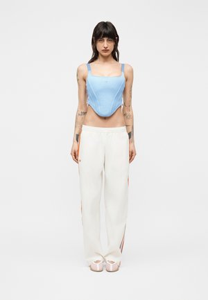 Femeie în picioare, purtând un corset din plasă albastru deschis, pantaloni largi albi cu dungi portocalii și sandale bej, cu tatuaje pe brațe.