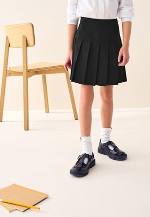 Kind in zwarte geplooide rok, witte sokken en zwarte schoenen staat bij een houten stoel en notitieboekjes met potlood op de vloer.