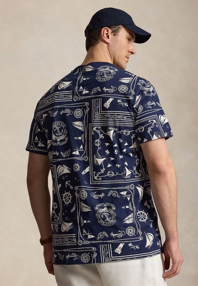 Uomo che indossa un berretto blu navy e una camicia blu navy a maniche corte con stampa nautica bianca, inclusi ancore, navi, timoni e fari, girato di spalle.