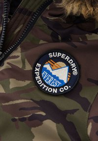 Kameru jaka audums ar apaļu izšūtu uzšuvi, uz kuras attēlota kalns un uzraksts "SUPERDRY EXPEDITION CO." zilā, oranžā un baltā krāsā.