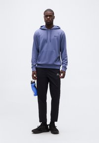 Homme portant un sweat à capuche bleu Nike et un pantalon noir Nike, debout et tenant une bouteille d'eau bleue devant un fond blanc uni.