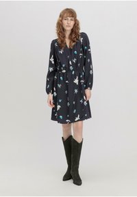 Dunkelblumenkleid mit langen  Ärmeln, elastischem Bund und knielangem Saum, das farbenfrohe Blumenmuster auf navyfarbenem Hintergrund zeigt. Kombiniert mit hohen, dunklen Stiefeln.