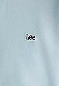 Jasnoniebieska bawełniana tkanina o teksturowanym wykończeniu z haftowanym białym i granatowym logo "Lee". Minimalistyczny design, bez dodatkowych wzorów.
