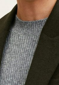 Donkergroene blazer met een kraag die een lichtgrijs ribgebreid sweater ontmoet. De materialen lijken zacht, met een gestructureerd breipatroon op de sweater.