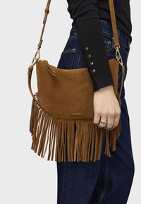 Borsa a tracolla in suede marrone con dettagli in frange, componenti dorati e tracolla regolabile, indossata su una mano, abbinata a denim scuro.