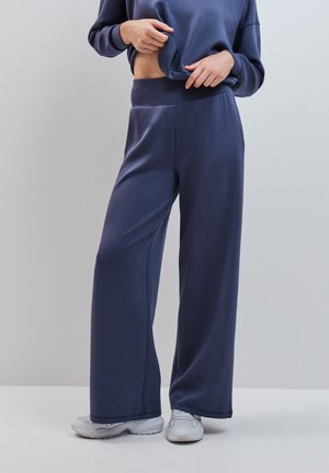 Personne portant un pantalon large bleu foncé et un sweat assorti, dévoilant une petite partie du ventre, associé à des baskets blanches.