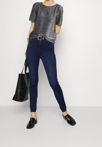 Sprankelende grijze top met korte mouwen, hoog opgesneden donkerblauwe jeans, zwarte riem, zwarte getextureerde instappers en een zwarte schoudertas.