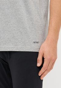 Šedé tričko s krátkým rukávem vyrobené z bavlněné směsi, s prošívaným lemem a malou černou nášivkou "Carhartt" na dolní levé straně.