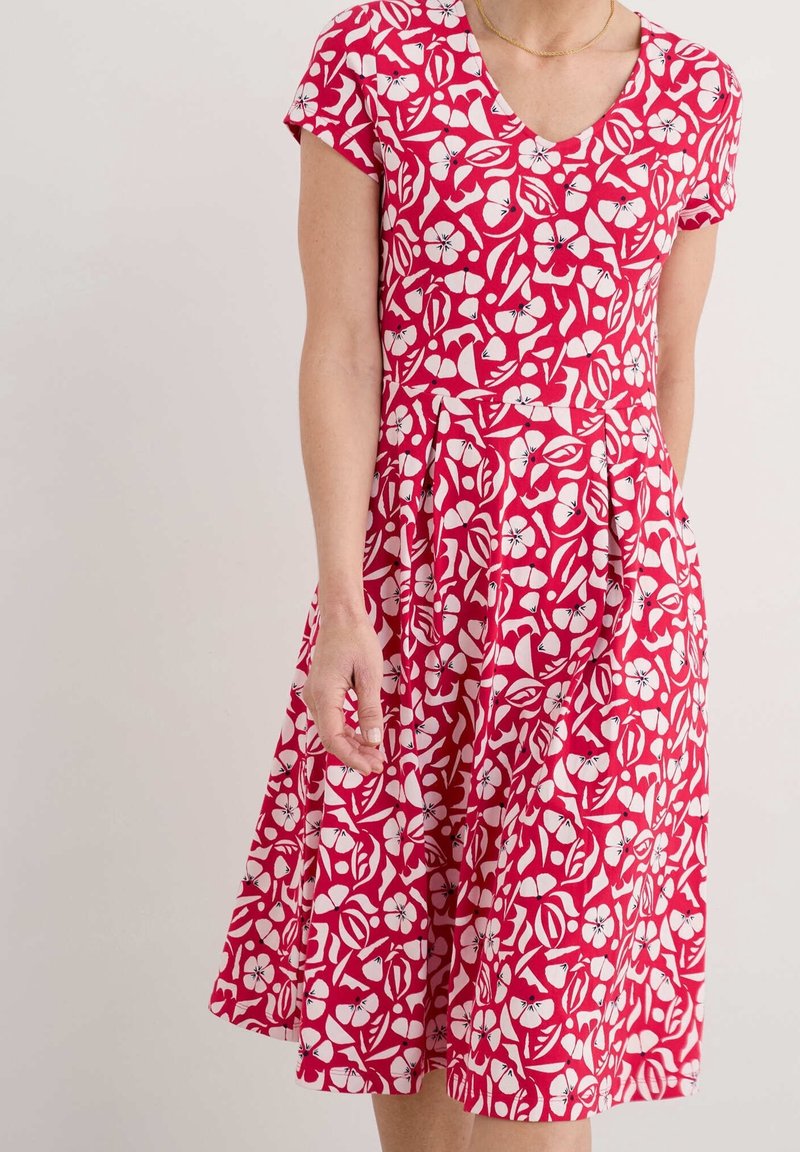 Femme portant une robe rouge avec des motifs floraux et de feuilles blancs, manches courtes et jupe plissée, debout devant un fond clair uni.