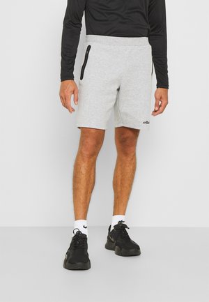 Mand iført lysegrå shorts med sorte lynlåslommer, sort langærmet top, hvide Nike-sokker og sorte sportssko, der står på en grå gulv.