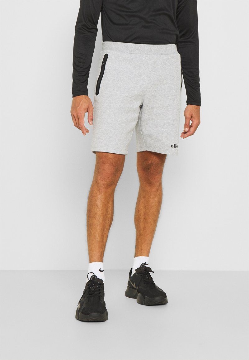 Homme portant un short gris clair avec des poches zippées noires, un haut à manches longues noir, des chaussettes blanches Nike et des chaussures de sport noires, debout sur un sol gris.
