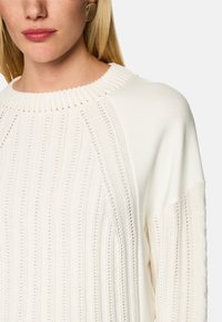 Cremefarbener Pullover mit geripptem Muster, einer weiten Rundhalslinie und weichem Material, akzentuiert durch horizontale und vertikale Muster.