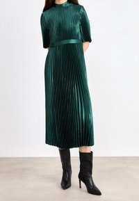 Groene plissé midi-jurk met korte mouwen en een satijnen tailleband, gemaakt van glanzende, gestructureerde stof. Gepaard met zwarte enkellaarsjes met hak.