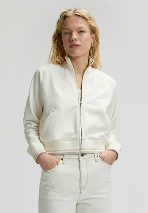 Kobieta o kręconych blond włosach, ubrana w kremową kurtkę na zamek oraz pasujące wysokie spodnie z rękami w kieszeniach.