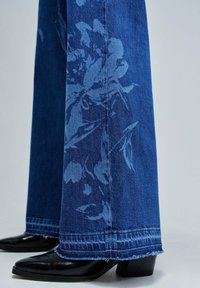 Jeans de mezclilla de pierna ancha en azul oscuro, con un sutil patrón floral en un azul más claro. Bajo recortado con bordes deshilachados y elegantes zapatos de tacón negros.