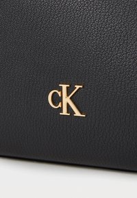 Black textured leather handbag featuring a gold "cK" logo emblem. The surface is embossed with a subtle grain pattern.