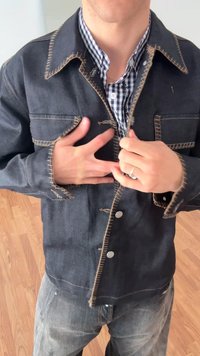 Casaco de ganga escura com costuras contrastantes em castanho claro, fecho de botões e bolsos no peito. Usado sobre uma camisa às riscas, com um corte descontraído.