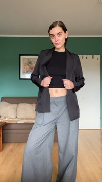 Μαύρο σακάκι προσαρμοσμένο πάνω από κοντή μαύρη μπλούζα, συνδυασμένο με ανοιχτό γκρι παντελόνι με φαρδιά ποδαρικά. Ο ιστός φαίνεται λείος με μια ελαφριά γυαλάδα.