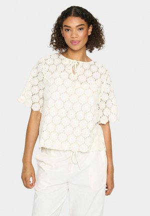 Top in pizzo color avorio con un design delle maniche a scallop e un colletto con fiocco, caratterizzato da un motivo floreale in tutto. Abbinato a pantaloni chiari.