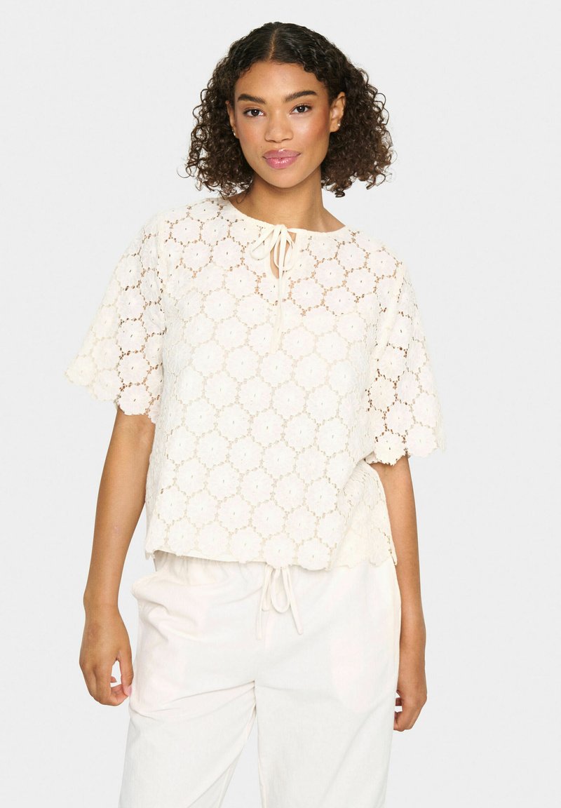 Top in pizzo color avorio con un design delle maniche a scallop e un colletto con fiocco, caratterizzato da un motivo floreale in tutto. Abbinato a pantaloni chiari.