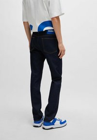 Jeans de mezclilla azul oscuro con un corte recto, bolsillos traseros y costuras naranjas, combinados con zapatillas blancas y azules y una camiseta gráfica blanca.