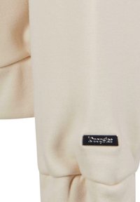 Beige sweatshirt featuring a soft texture, ribbed cuffs, and a small metallic brand label on the sleeve.