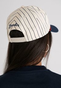 Hvit caps med marineblå striper, "Berlin" brodert i marineblått. Justerbar stropp bak, marineblå skygge, teksturert stoff.