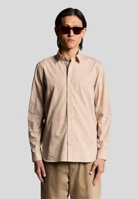 Långärmad ljusbeige skjorta med subtila blommönster, knappad krage och vita knappar, kombinerad med khakifärgade byxor och mörka solglasögon.
