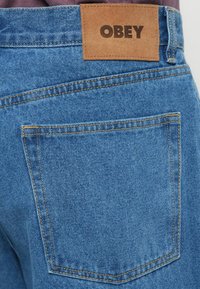Modre denim hlače s rjavim usnjenim zakrpom z odtisnjenim napisom "OBEY". Vidno šivanje in podrobnosti na zadnjem žepu obogatita dizajn.