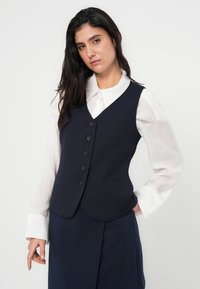 Μπλε σκούρο ριμπ μπλουζάκι χωρίς μανίκια με V λαιμόκοψη, πέντε κουμπιά και πλαϊνές τσέπες, φορεμένο πάνω από λευκό πουκάμισο με μακριά μανίκια και μανσέτες με κουμπιά.