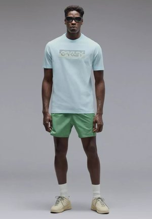 Homme portant des lunettes de soleil noires, un t-shirt Oakley bleu clair, un short vert, des chaussettes blanches et des baskets beiges, debout devant un fond gris.