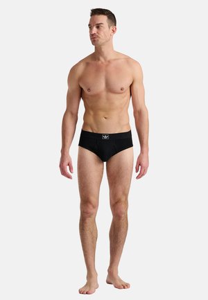 Shirtless man standing barefoot wearing black briefs with a logo on the waistband, looking to the left against a plain white background.