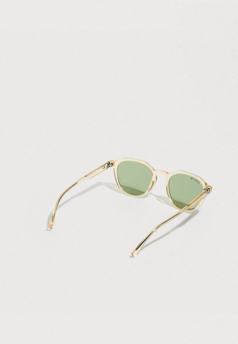 Lunettes de soleil à monture beige transparente avec des verres ronds verts, posées ouvertes sur une surface blanche.