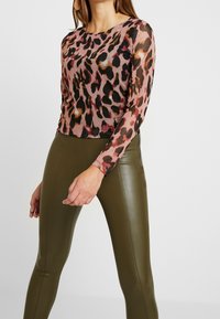 Langärmliges Oberteil aus durchscheinendem, leichtem Stoff mit einem pinken und schwarzen Leopardenmuster, kombiniert mit olivgrünen, hochtaillierten Leggings.