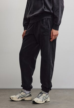 Personne portant un pantalon de survêtement noir, un sweat-shirt noir assorti, des chaussettes blanches jusqu'aux chevilles et des baskets blanches et grises, debout sur un sol gris.