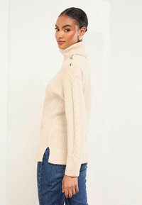 Bežový rolák s pletenými detailmi pozdĺž rukávov a zlatými gombíkovými akcentmi. Bočné rozparky na spodnom leme pre väčšiu pohyblivosť.