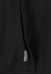 Schwarzes, strukturiertes Stoffkleidungsstück mit Knopfverschluss, das ein Etikett mit der Aufschrift "SUBLEVEL" nahe der unteren Kante trägt. Sauberes, minimalistisches Design.
