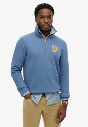Homme portant un sweat-shirt bleu à zip quart avec un logo jaune sur une chemise rayée à boutons, pantalon beige et montre-bracelet, debout avec une main dans la poche.