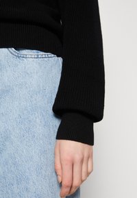 Schwarzer gerippter Strickpullover mit weitem Bündchen, kombiniert mit hellblauen Jeans, die einen strukturierten Stoff und einen lässigen Schnitt zeigen.